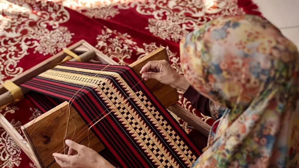 Foto pengrajin tapis sedang menapis kain tapis menggunakan alat peraga tradisional di workshop, menampilkan keahlian dan dedikasi dalam pembuatan produk berkualitas.